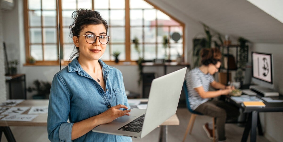 professional woman in office holding laptop, colleague in the background working at desk